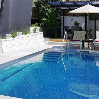 Ein schöner Pool mit klarem Wasser in einem modernen Garten. Im Hintergrund sind Liegestühle und ein Sonnenschirm zu sehen.