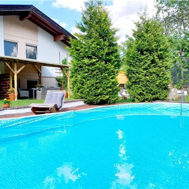 Ein klarer Pool umgeben von grünen Bäumen und einem gemütlichen Garten. Im Hintergrund sieht man ein modernes Haus mit einer überdachten Terrasse.