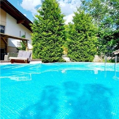 Ein klarer Pool umgeben von grünen Bäumen und einem modernen Haus. Die Sonne scheint und der Himmel ist blau mit einigen Wolken.