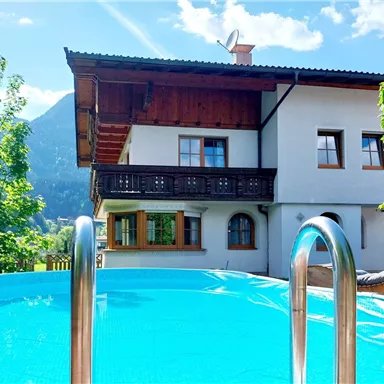 Ein schönes Haus mit einem Pool und einer Berglandschaft im Hintergrund. Die Sonne scheint und es gibt viel Grün rund um das Gebäude.