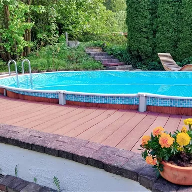 Ein sauberer Pool mit klarem Wasser ist von einem Holzdeck umgeben. Im Hintergrund befinden sich grüne Bäume und eine bepflanzte Ecke.