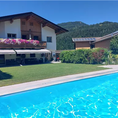 A beautiful house with a pool and a well-maintained garden. The surroundings are filled with mountains and sunlight.