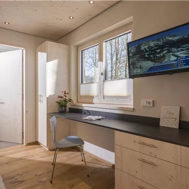 A modern bedroom with wooden flooring and clean lines. There is a desk, a television, and large windows that let in plenty of light.