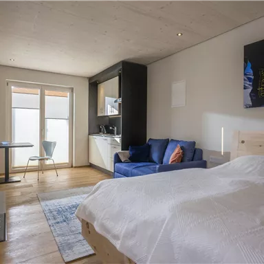 A bright room with a comfortable bed, a blue sofa, and a small dining table. 

Large windows provide plenty of natural light.