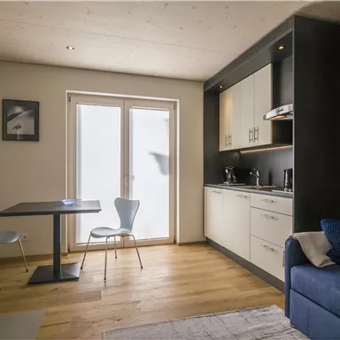 A modern kitchen with a table and two chairs. The room is bright and inviting, with a cozy sofa and large windows.
