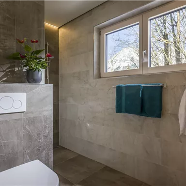 A modern bathroom with a toilet, towels, and a window that provides light and a view of trees. The walls are in neutral colors.