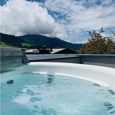 Ein Whirlpool auf einer Terrasse mit Blick auf grüne Hügel und Berge. Der Himmel ist klar mit einigen Wolken.