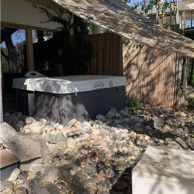 Ein Whirlpool steht auf einem Bett aus Steinen, umgeben von Pflanzen und Holz. Im Hintergrund sind sonnige Bäume und eine hell beleuchtete Terrasse zu sehen.
