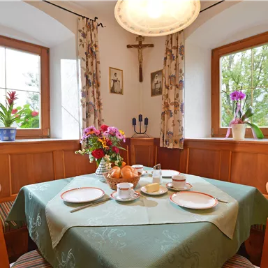 Ein einladender Essbereich mit einem Tisch, der mit Geschirr und einer Blumenmitte dekoriert ist. Die Fenster bieten einen Blick ins Grüne und das Zimmer hat eine warme, gemütliche Atmosphäre.