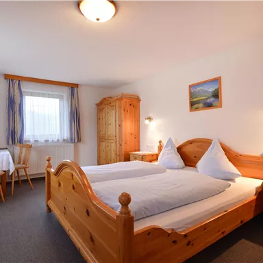 Ein gemütliches Zimmer mit zwei Betten aus Holz und blauem Dekor. Im Hintergrund sind ein Tisch, Stühle und ein kleiner Fernseher zu sehen.