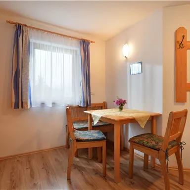 Ein gemütlicher Essbereich mit einem Tisch und zwei Stühlen. Fenster mit Vorhängen und eine Blume auf dem Tisch sorgen für eine einladende Atmosphäre.