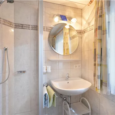 Ein modernes Badezimmer mit einer Dusche, einem Waschbecken und einem runden Spiegel. Große Fenster sorgen für viel Tageslicht.