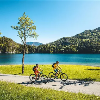 Two children are riding bicycles along the shore of a clear lake. The landscape is green and surrounded by hills, under a bright blue sky.