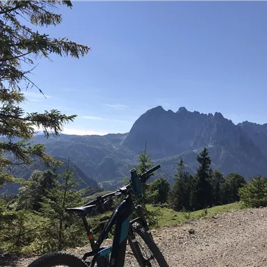 A mountain bike stands on a gravel path with a view of the mountains. The sky is clear and the landscape is green and hilly.