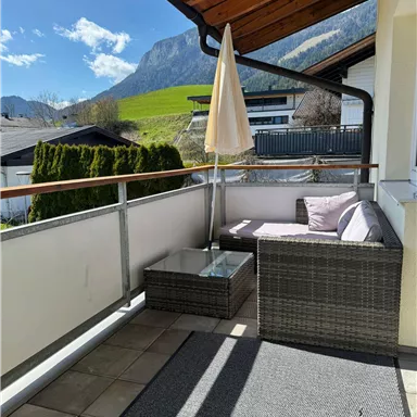 A balcony with a cozy lounge area and a sunshade. In the background, green meadows and mountains can be seen.