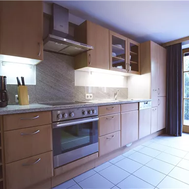 Eine moderne Küche mit Holzschränken und einer Edelstahl-Spüle. Große Fenster sorgen für viel Tageslicht.