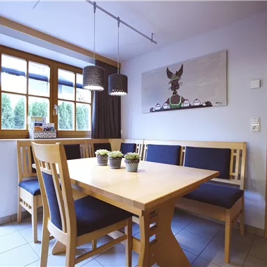 Ein heller Essbereich mit einem Holzstisch und gemütlichen Bänken. Große Fenster lassen viel Tageslicht herein.