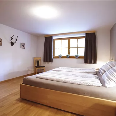 Ein modernes Schlafzimmer mit einem Doppelbett und Holzfußboden. Die Fenster bieten Tageslicht und eine gemütliche Atmosphäre.