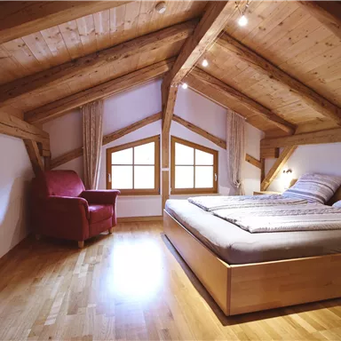 Ein gemütliches Dachzimmer mit einem großen Bett und einem roten Sessel. Die Holzdecken und der Boden verleihen dem Raum eine warme Atmosphäre.