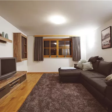 Ein modernes Wohnzimmer mit einem bequemen schwarzen Sofa und einem Fernseher. Große Fenster sorgen für viel Tageslicht und die Wände sind mit Bildern dekoriert.
