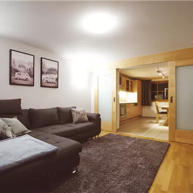 Ein modernes Wohnzimmer mit einem großen, komfortablen Sofa und einem Teppich. Im Hintergrund ist die Küche sichtbar, ergänzt durch einige stilvolle Wandbilder.