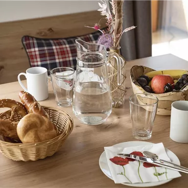 A beautifully set table with fresh rolls, fruit, and glasses. In the background, there is a carafe of water and a flower.