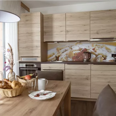 A modern kitchen with wooden cabinets and a dining area. On the table, there is fresh bread, dishes, and a glass of water.