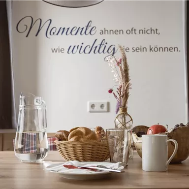 A cozy table with a fruit basket, rolls, and a coffee cup. In the background, a quote is hanging on the wall.