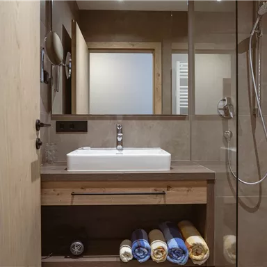 A modern bathroom with a sink and a shower. Towels are neatly stacked in the cupboard.