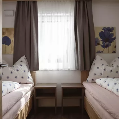 A simple, bright room with two single beds and a large window front. The walls are decorated with colorful pictures, and curtains create a cozy atmosphere.