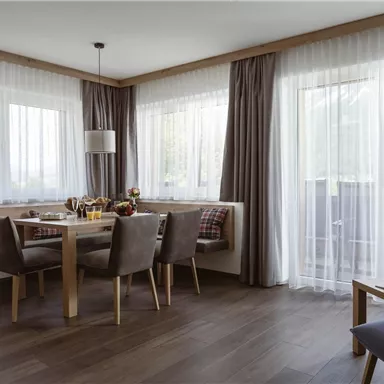 A modern dining room with a wooden table and cozy chairs. Large windows let in plenty of daylight and offer a view outside.