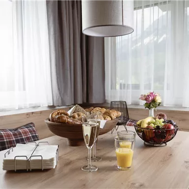 An inviting breakfast table with fresh pastries, fruit, and glasses. The room has large windows with curtains and offers a cozy atmosphere.