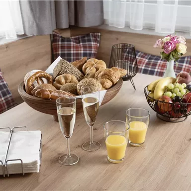A set table with a selection of fresh pastries, fruits, and drinks. In the background, cozy cushions can be seen.