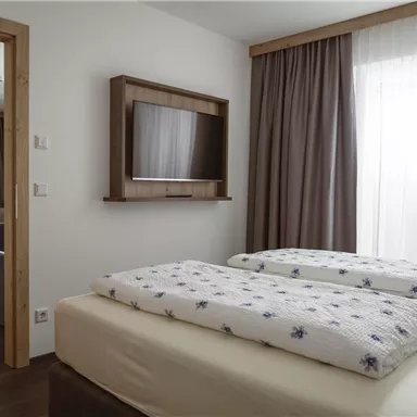 A cozy bedroom with two beds and a television on the wall. The windows are covered with curtains and let in a lot of light.