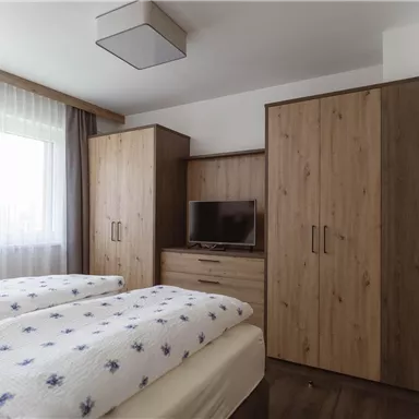 A cozy bedroom with two single beds, a wooden wardrobe, and a television. The window provides plenty of daylight.