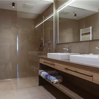 A modern bathroom with a spacious shower and two sinks. The walls are in neutral tones and the furniture is made of wood.