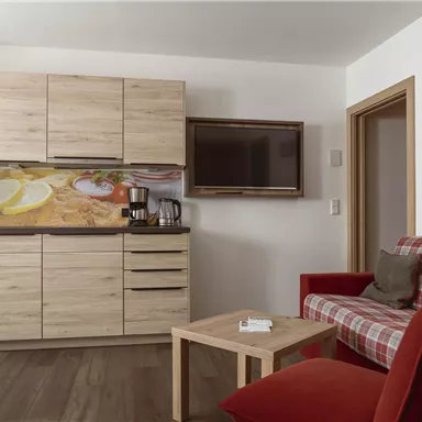 A modern kitchen with wooden furniture and a television on the wall. In the foreground is a red sofa and a small coffee table.