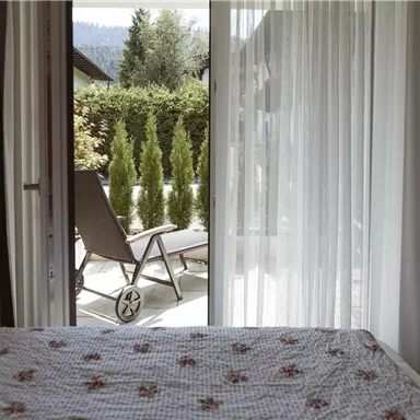 A cozy bedroom with a view of a terrace. In the background, green plants and a lounge chair can be seen.