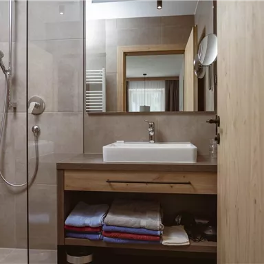 A modern bathroom with a shower and a sink. Towels are neatly stacked on a wooden shelf.