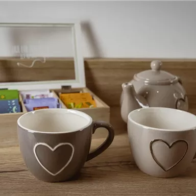Two decorative cups with hearts and a teapot are on a wooden surface. In the background, there is a box with various tea bags.