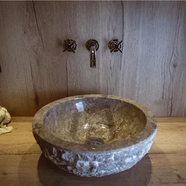 An elegant stone sink on a wooden surface. Stylish faucets are visible in the background.
