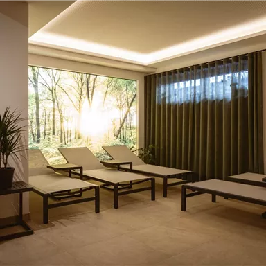 A relaxing room with loungers and a large window that shows a forest with sunlight. The atmosphere is calm and inviting.
