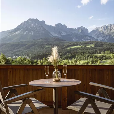 A cozy balcony with a round table and two chairs. In the background, majestic mountains and a clear sky can be seen.