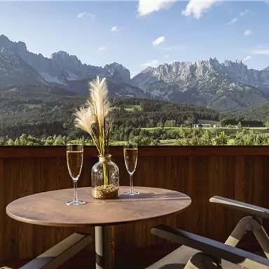 A table on a balcony with two glasses and a decorative vase. In the background, impressive mountains and a picturesque landscape can be seen.