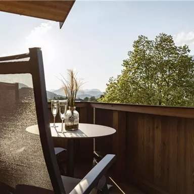 A cozy balcony with a chair and a small table. In the background, trees and mountains can be seen under a clear sky.