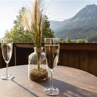 A beautiful table with two glasses and a vase on a balcony. In the background, majestic mountains and a clear sky can be seen.