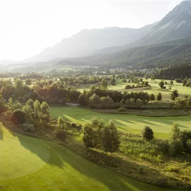 A wide golf course surrounded by gentle hills and trees. In the background, mountains and a clear sky can be seen.