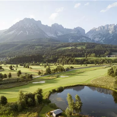 A picturesque golf course with a clear pond and gentle hills. In the background, majestic mountains rise under a blue sky.