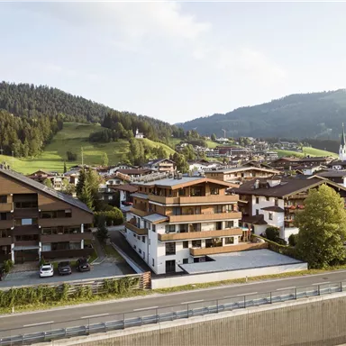A picturesque view of a village with modern and traditional buildings, surrounded by gentle hills. The sky is clear and the landscape is green.