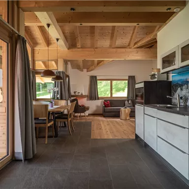 A modern kitchen with wooden ceilings and large windows. The space is open and bright, with a dining area and a cozy living room.